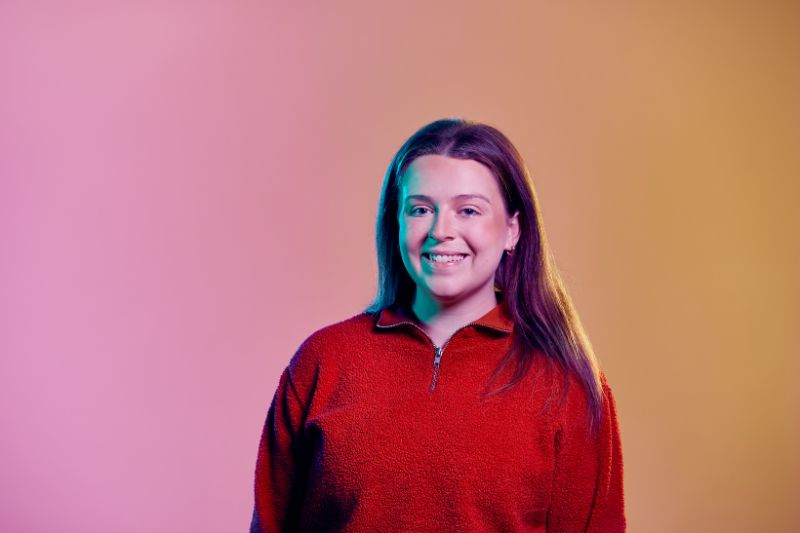 Dark haired female wearing a red jumper standing in fron t of pink orange ombre coloured background